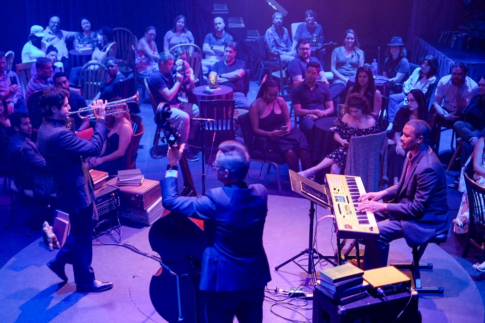 Bathed in purple light, musicians play trumpet, bass and keyboard on stage in front of an audience.
