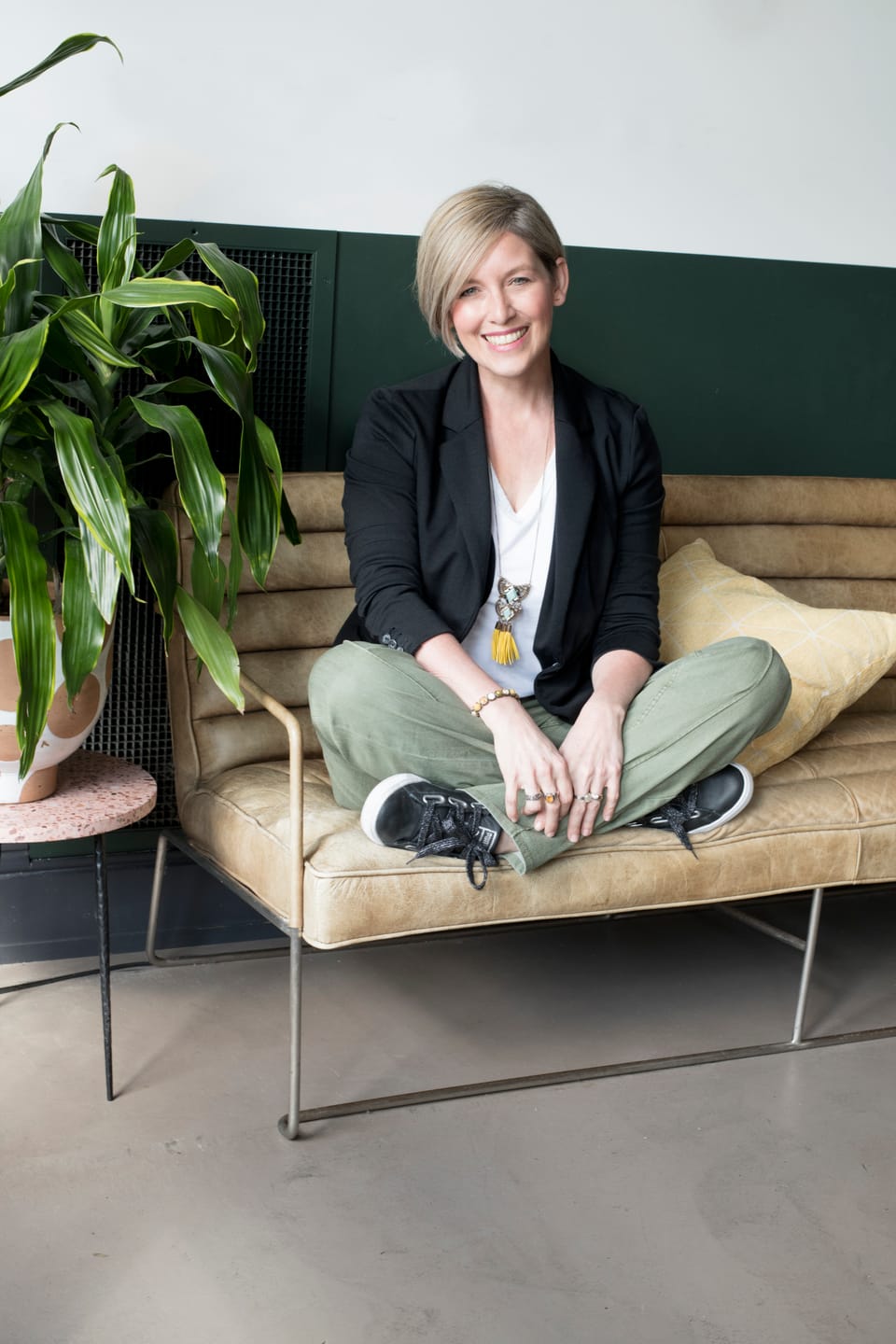 A woman sits cross-legged on a couch. A potted plant is next to her. 